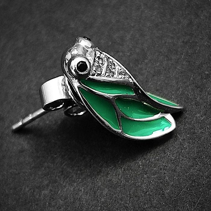 Paire de boucles d’oreilles cigale argent 925, ailes vert émail, posées sur surface noire.